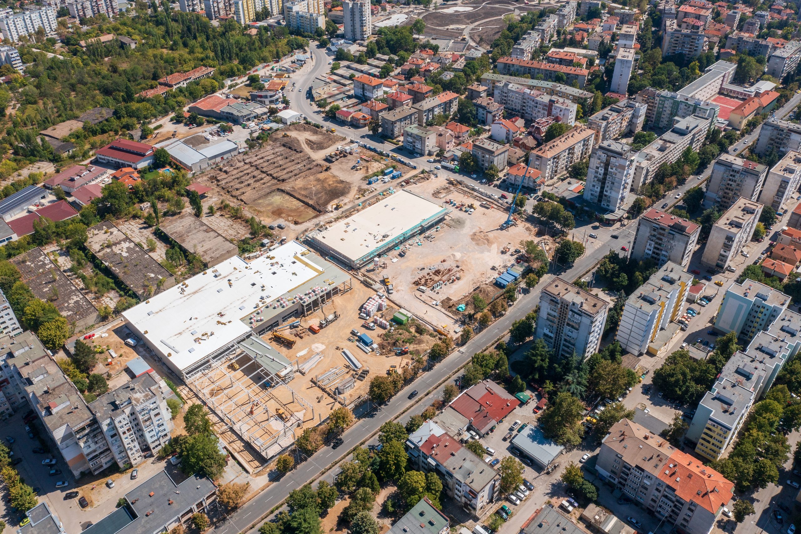 Aerial view of a large construction site with excavators and materials, separated by a street from occupied apartment blocks and houses on every side